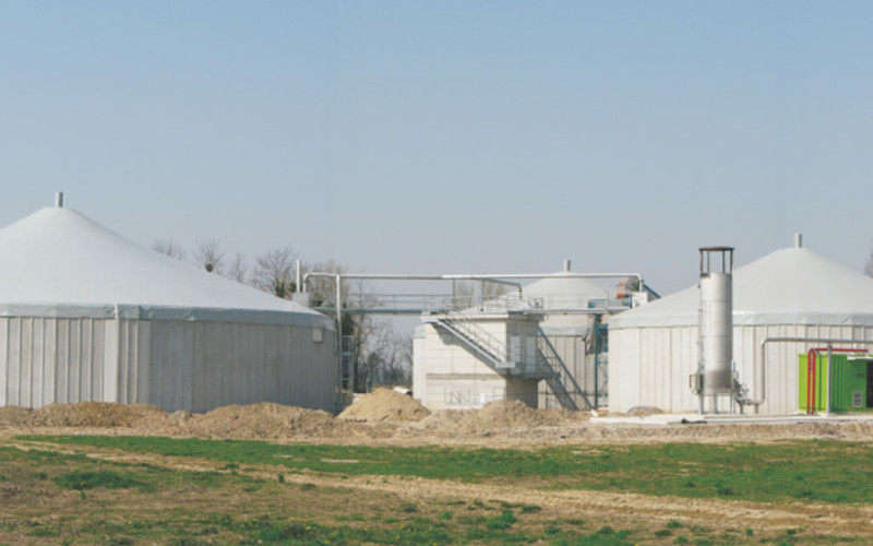 Impianto di biogas con grandi serbatoi circolari coperti da teli di protezione, tubazioni e strutture metalliche per il trasporto del gas.