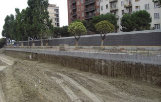 Cantiere urbano con scavo profondo in primo piano e barriera fonoassorbente in cemento sullo sfondo.