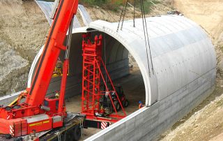 Fase di costruzione di una galleria stradale con elementi prefabbricati in cemento. Una grande gru mobile rossa sta sollevando una sezione ad arco del tunnel per posizionarla in loco. Operai e attrezzature di supporto sono presenti nel cantiere, con pareti di contenimento in cemento ai lati e un'area di scavo coperta con teli di plastica.