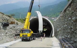 Montaggio di un elemento prefabbricato in cemento per la costruzione di una galleria stradale. Una gru gialla solleva e posiziona un grande arco all'ingresso del tunnel, mentre operai con caschi di sicurezza lavorano all'interno.