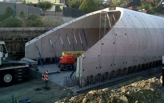 Costruzione di una galleria prefabbricata in cemento. Una grande struttura ad arco è in fase di assemblaggio, sostenuta da puntelli metallici. Una gru della ditta Casella Autogru è presente sul cantiere, insieme a un sollevatore telescopico rosso.