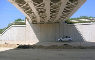 Vista sotto un ponte con struttura in cemento armato e travatura reticolare metallica.