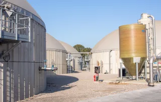 Primo piano di un impianto di biogas con biodigestori coperti, tubazioni industriali, valvole e una cisterna verticale per il trattamento dei gas.