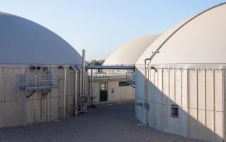 Primo piano di un impianto di biogas con biodigestori coperti da cupole flessibili, tubazioni di collegamento e passerelle metalliche per l’accesso alla manutenzione.