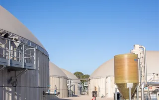 Primo piano di un impianto di biogas con biodigestori coperti, tubazioni industriali, valvole e una cisterna verticale per il trattamento dei gas.