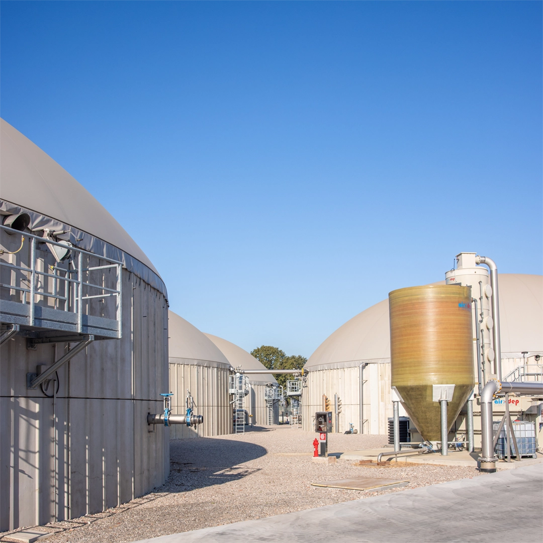 Primo piano di un impianto di biogas con biodigestori coperti, tubazioni industriali, valvole e una cisterna verticale per il trattamento dei gas.