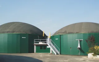 Due grandi biodigestori agricoli di colore verde con coperture a cupola nera, utilizzati per la produzione di biogas. Una scala metallica conduce a una piattaforma di accesso tra i due serbatoi. Sul lato destro, è visibile un sistema di miscelazione montato sulla parete.