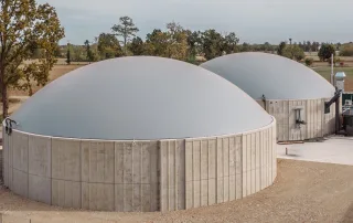 Due biodigestori agricoli cilindrici in cemento con copertura a cupola grigia. ul lato destro dell'immagine è visibile un sistema di tubazioni e macchinari collegati all'impianto.