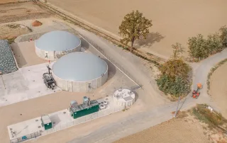Veduta aerea di un impianto di biodigestione anaerobica in un'area agricola, con due grandi serbatoi cilindrici in cemento dotati di copertura a cupola.