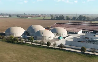 Vista aerea di un impianto di biogas con diversi biodigestori a cupola situato in un'azienda agricola. Sullo sfondo si vedono campi coltivati, edifici agricoli con tetti in metallo e recinti per il trattamento di materiali organici.