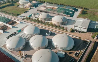 Veduta aerea di un impianto di biogas con più biodigestori coperti, aree di stoccaggio per biomasse e strutture di supporto.