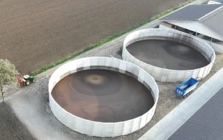 Vista aerea di due grandi vasche circolari in cemento utilizzate per la raccolta e lo stoccaggio di liquami agricoli, situate accanto a un capannone e circondate da campi coltivati.