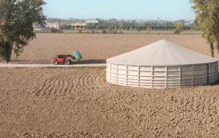 Vista panoramica di una vasca agricola circolare con copertura in telo, situata in un campo coltivato. Un trattore rosso con un caricatore frontale trasporta una balla di fieno lungo una strada sterrata accanto alla vasca.