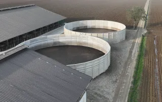 Vista aerea di due grandi vasche agricole circolari in calcestruzzo, situate accanto a una stalla coperta in un'azienda agricola. Intorno si estendono campi coltivati e una strada sterrata.
