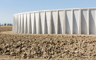 Vasca agricola in cemento con struttura circolare e pareti esterne rinforzate da scanalature verticali. La vasca è posizionata su un terreno agricolo arato.