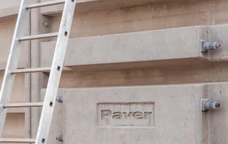 Dettaglio di una parete in cemento armato di una vasca agricola, con il logo "Paver" inciso sulla superficie. Una scala in alluminio è appoggiata alla struttura.