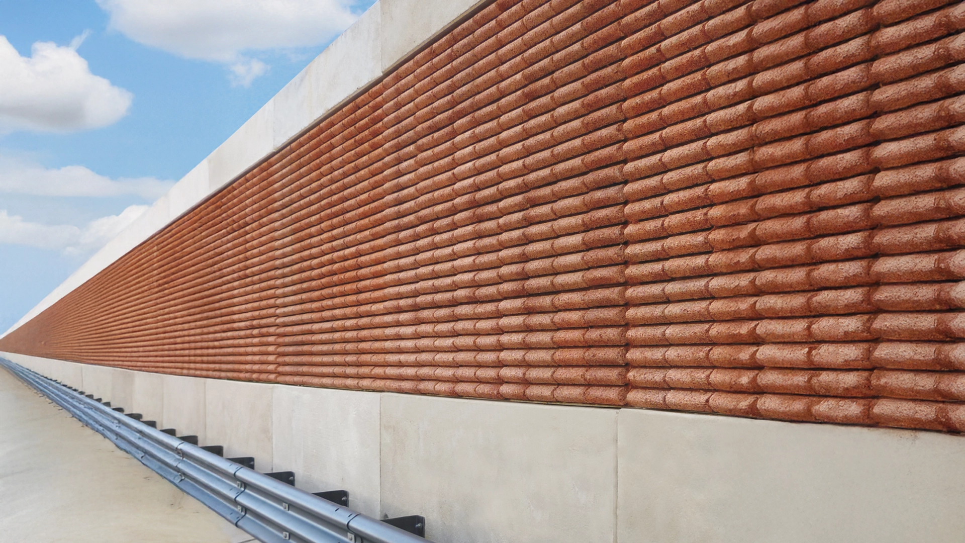 Primo piano di una barriera fonoassorbente lungo una strada, caratterizzata da una texture in rilievo con elementi in laterizio e una base in cemento. A fianco, un guardrail metallico segue il bordo della carreggiata sotto un cielo azzurro con nuvole sparse.