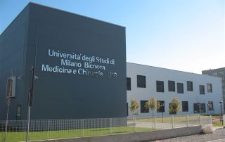 Edificio prefabbricato dell'Università degli Studi di Milano Bicocca, Medicina e Chirurgia U18, con facciata moderna in pannelli grigi e bianchi, finestre nere incassate e recinzione metallica.