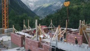 Cantiere in una valle montana con murature in blocchi di laterizio Bioplus in costruzione, con operai al lavoro e una gru in funzione.