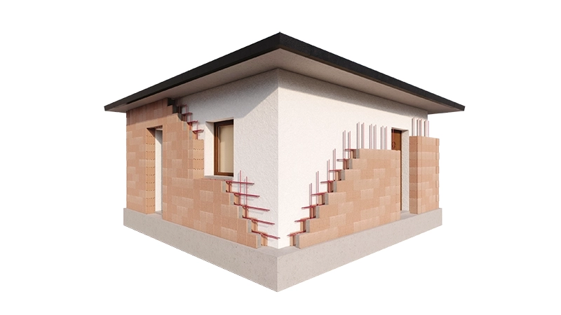 L'immagine mostra un edificio ad angolo con parte delle pareti esterne completate e altre in fase di costruzione. Alcune sezioni della muratura in laterizio sono esposte, mostrando barre di rinforzo metalliche e strati di isolamento. Il tetto è scuro e spiovente, mentre la parte completata della facciata è intonacata di bianco.