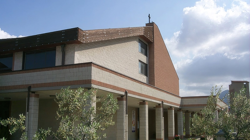 Una chiesa moderna con una struttura in mattoni e cemento, caratterizzata da un tetto spiovente con una croce sulla sommità. La facciata presenta una combinazione di superfici lisce e rivestimenti in mattoni rossi. Un portico con colonne rivestite in mattoncini chiari circonda l'ingresso