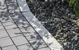 Cordolo in granito chiaro con finitura bocciardata, separa una pavimentazione in autobloccanti grigi da un'area decorata con ghiaia.