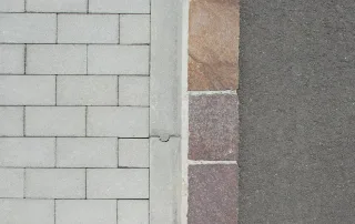 Cordolo stradale in cemento con incastro, delimitante una pavimentazione in autobloccanti grigi e una carreggiata asfaltata con fascia di pietra naturale.