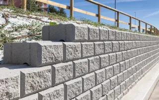 Muro di contenimento realizzato con blocchi Muricciolo grigi, installato lungo una strada con recinzione in legno e vegetazione sul pendio