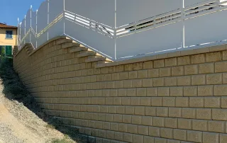 Muro di contenimento realizzato con blocchi Muricciolo color pietra alberese, completato da una recinzione bianca e un'elegante finitura superiore a gradoni.