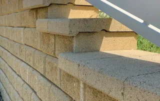 Dettaglio di un muro di contenimento in blocchi Muricciolo color pietra alberese, con finitura superiore a gradoni e recinzione bianca sovrastante.