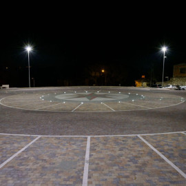 "Pavimentazione in pietra con motivo a rosa dei venti al centro, delimitata da linee bianche e illuminata da luci incassate.
