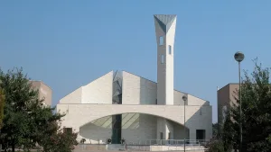 Una chiesa moderna con una struttura geometrica in pietra chiara, caratterizzata da un campanile alto e inclinato con aperture strette e verticali. La facciata presenta un ampio arco vetrato.