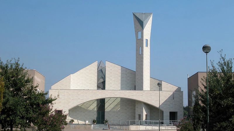 Una chiesa moderna con una struttura geometrica in pietra chiara, caratterizzata da un campanile alto e inclinato con aperture strette e verticali. La facciata presenta un ampio arco vetrato.