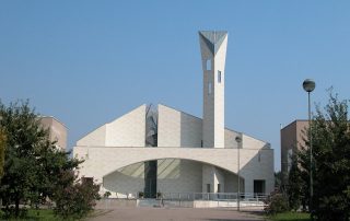 Una chiesa moderna con una struttura geometrica in pietra chiara, caratterizzata da un campanile alto e inclinato con aperture strette e verticali. La facciata presenta un ampio arco vetrato.