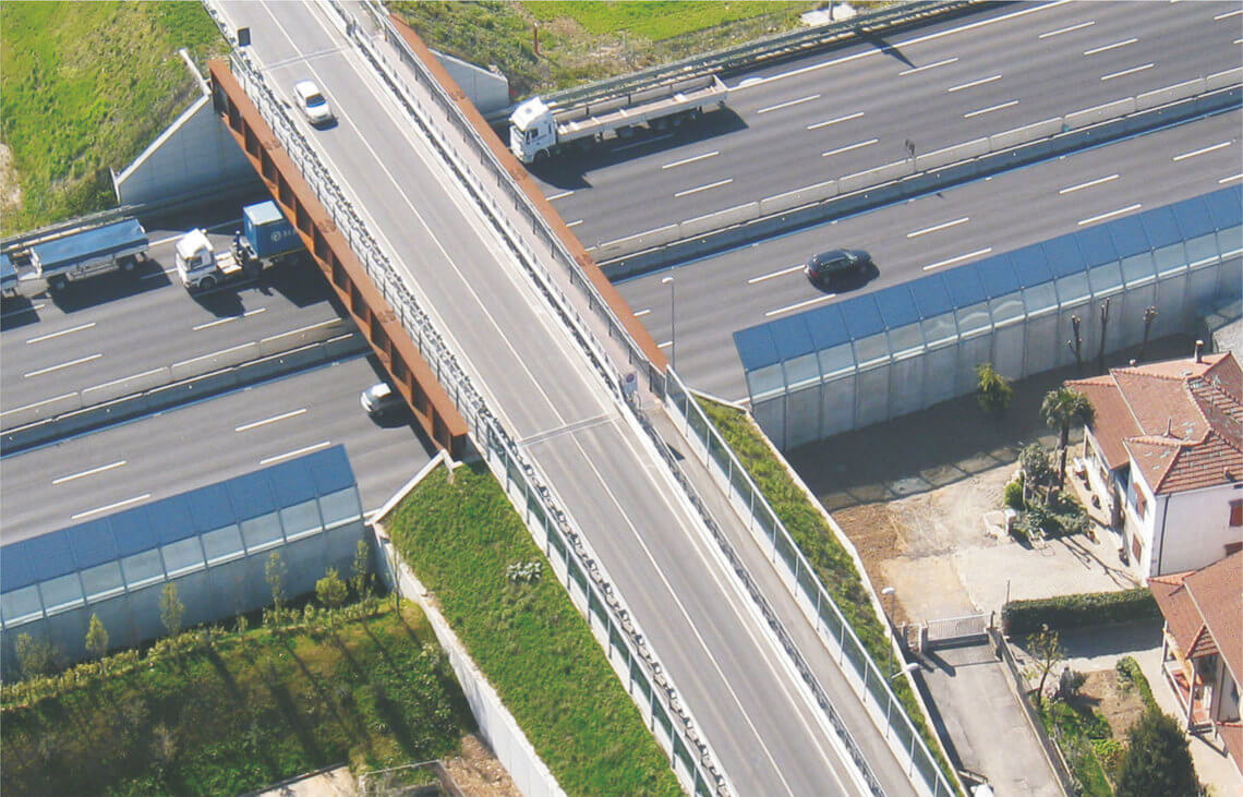 Vista aerea di un ponte stradale moderno in acciaio e cemento che attraversa un'autostrada a più corsie. La struttura include barriere di protezione trasparenti ai lati e pannelli fonoassorbenti lungo l'autostrada per ridurre il rumore.