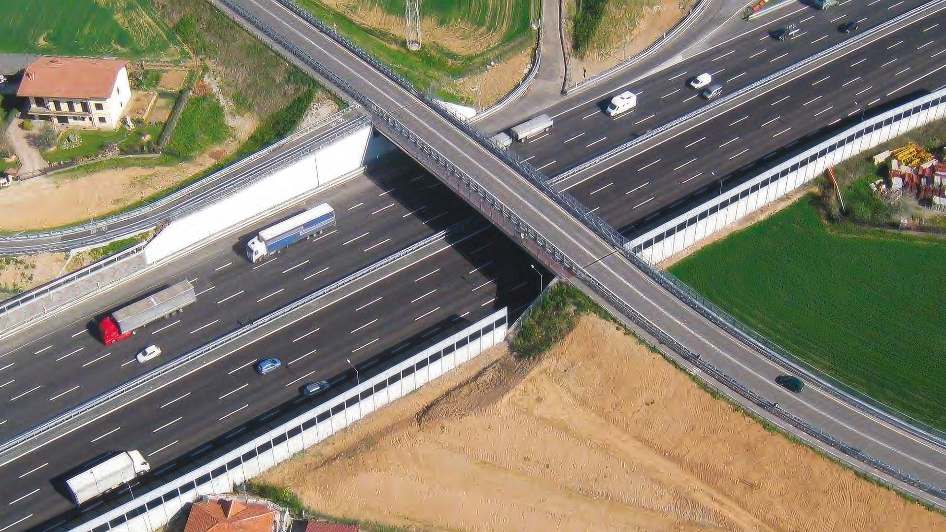 Veduta aerea di un'autostrada con più corsie e traffico di camion e automobili, attraversata da un cavalcavia stradale. Ai lati sono presenti aree di sosta, parcheggi e zone industriali.