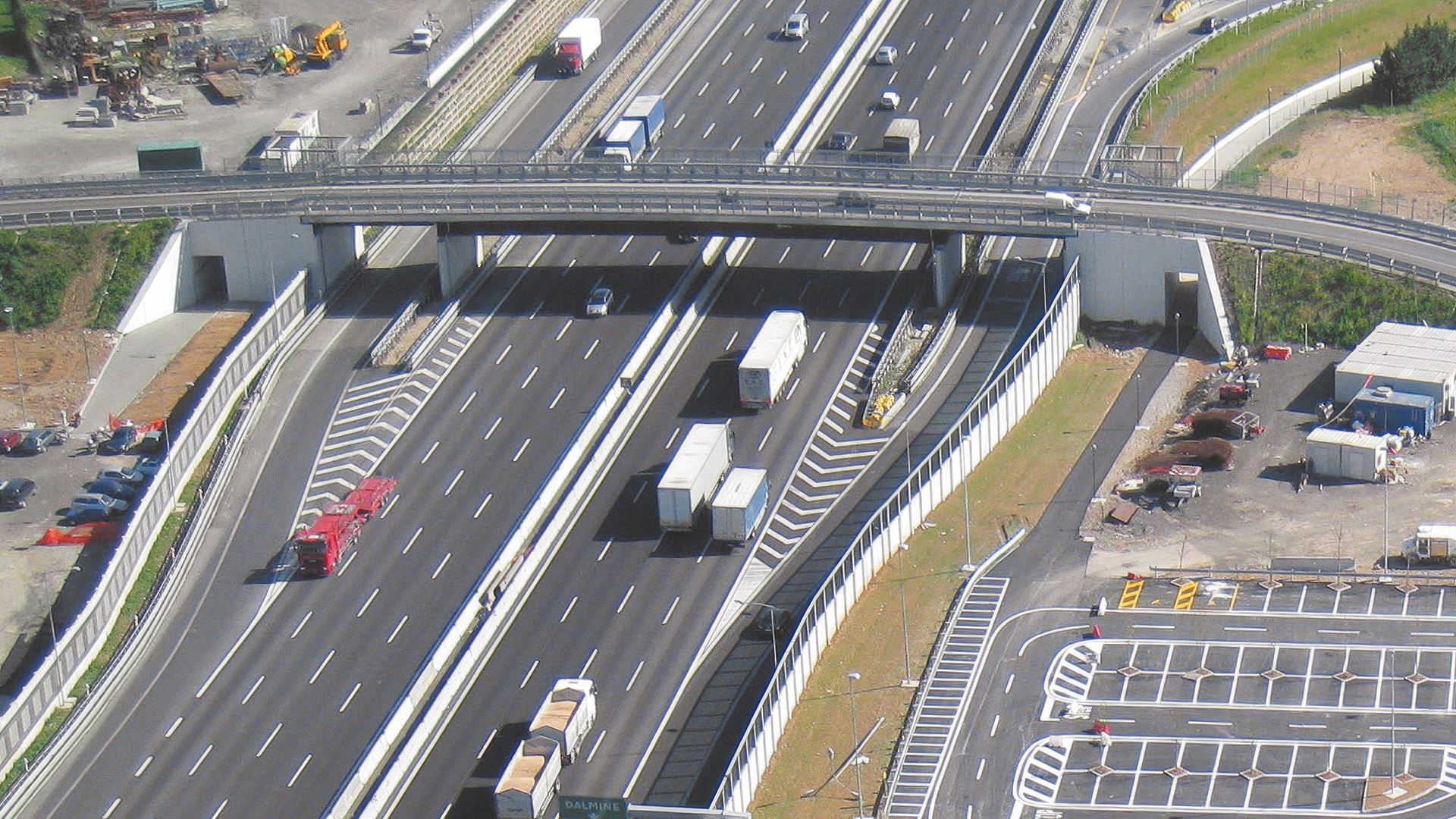 Veduta aerea di un'autostrada con più corsie e traffico di camion e automobili, attraversata da un cavalcavia stradale. Ai lati sono presenti aree di sosta, parcheggi e zone industriali.