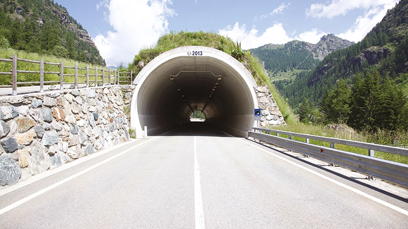 Strada di montagna che attraversa una galleria ad arco in calcestruzzo, circondata da rocce, vegetazione e paesaggio alpino.