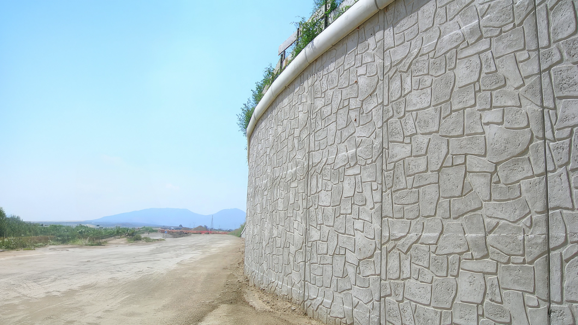 Muro di sostegno prefabbricato in calcestruzzo con finitura a finta pietra, installato lungo una strada sterrata, con vegetazione sulla sommità.