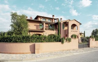 Villa in stile toscano con facciata in tonalità terracotta, persiane verdi e tetto in tegole, circondata da un giardino rigoglioso e una recinzione con cancello in ferro battuto.