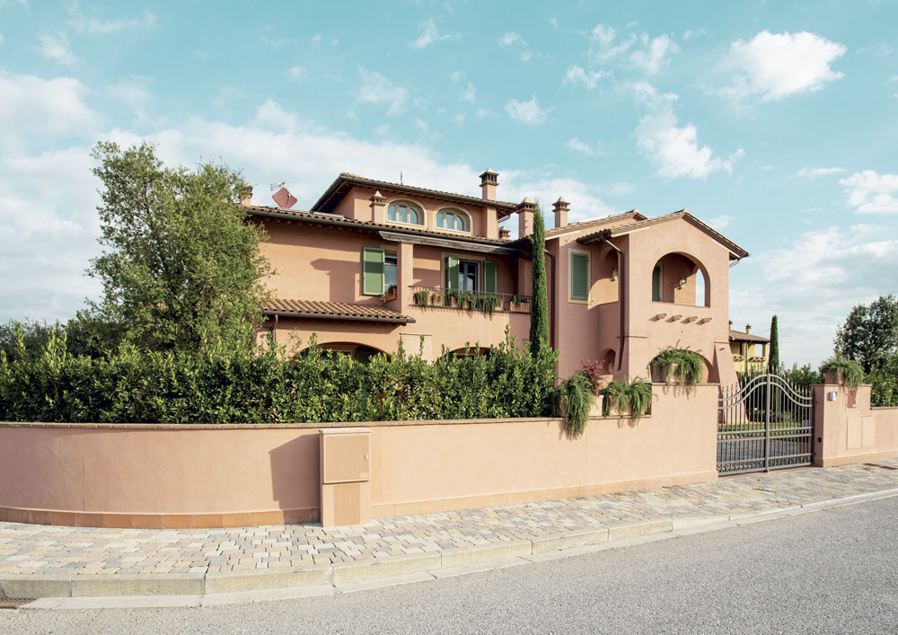 Villa in stile toscano con facciata in tonalità terracotta, persiane verdi e tetto in tegole, circondata da un giardino rigoglioso e una recinzione con cancello in ferro battuto.