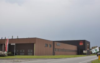 Edificio industriale della Musetti Caffè con design moderno e minimalista. La struttura è caratterizzata da una grande afacciata nera con un'imponente lettera "M" rossa. Il capannone principale presenta una struttura con tetto a shed e facciata marrone con il logo "Musetti".