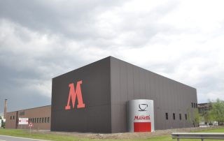 Edificio industriale della Musetti Caffè con design moderno e minimalista. La struttura è caratterizzata da una grande facciata nera con un'imponente lettera "M" rossa. Sulla destra, un cilindro grigio con il logo "Caffè Musetti Mio Espresso" evidenzia il marchio.