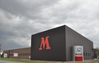 Edificio industriale della Musetti Caffè con design moderno e minimalista. La struttura è caratterizzata da una grande facciata nera con un'imponente lettera "M" rossa. Sulla destra, un cilindro grigio con il logo "Caffè Musetti Mio Espresso" evidenzia il marchio.