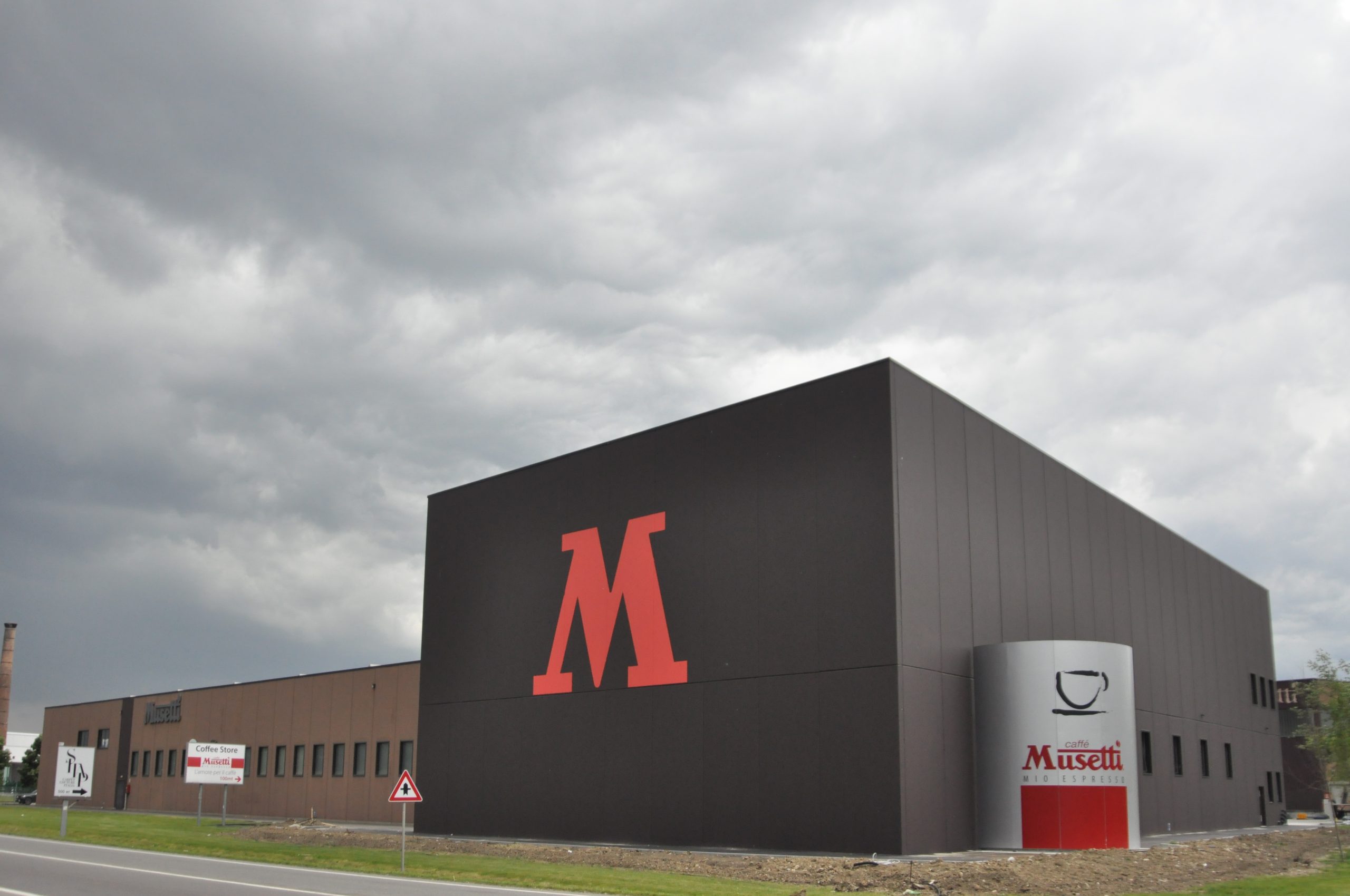 Edificio industriale della Musetti Caffè con design moderno e minimalista. La struttura è caratterizzata da una grande facciata nera con un'imponente lettera "M" rossa. Sulla destra, un cilindro grigio con il logo "Caffè Musetti Mio Espresso" evidenzia il marchio.