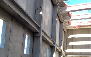 Interno di un edificio industriale prefabbricato in costruzione, con pareti in cemento, travi a vista e reti di sicurezza sulla copertura.