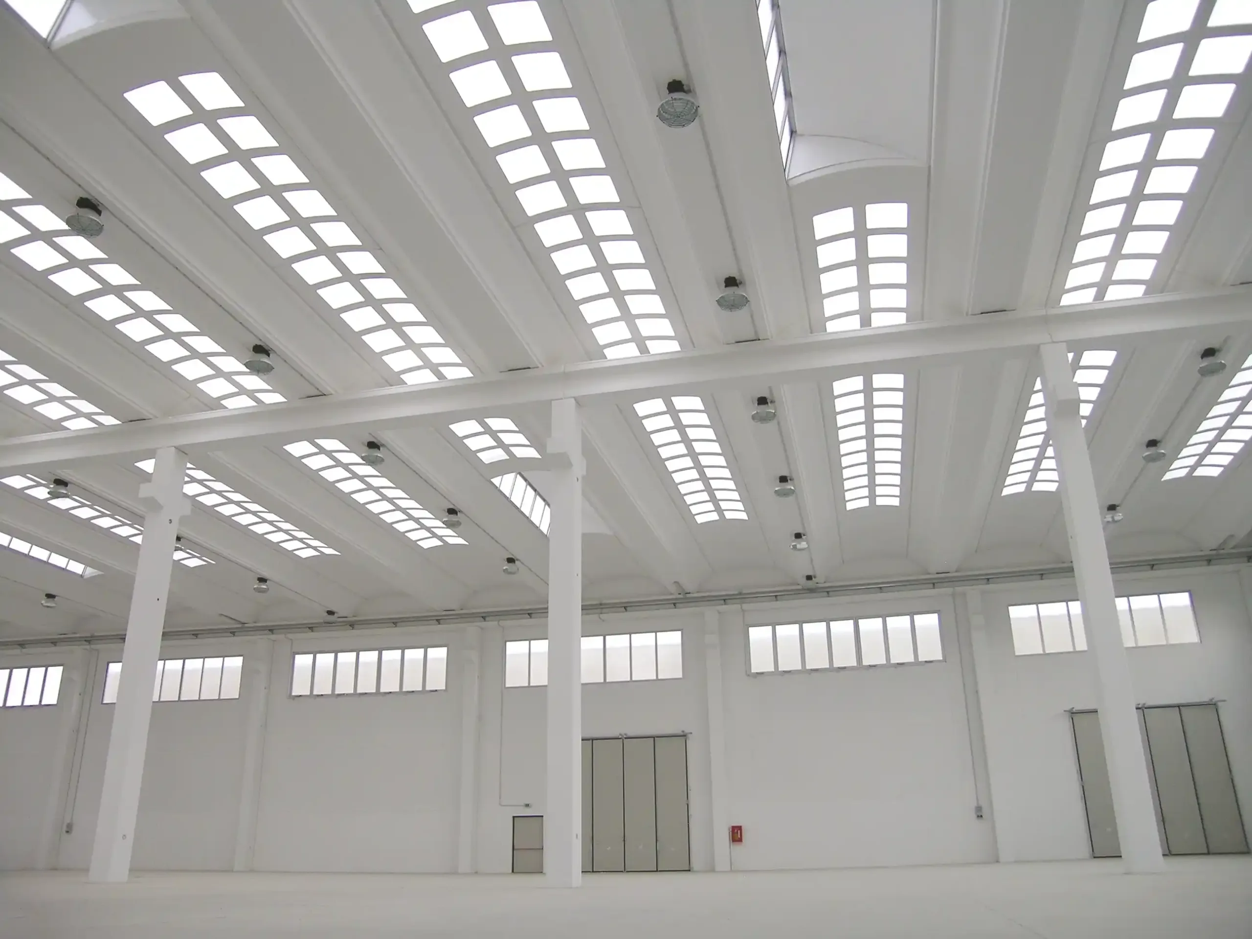 Interno di un capannone industriale con lucernari a soffitto e struttura in cemento bianco.