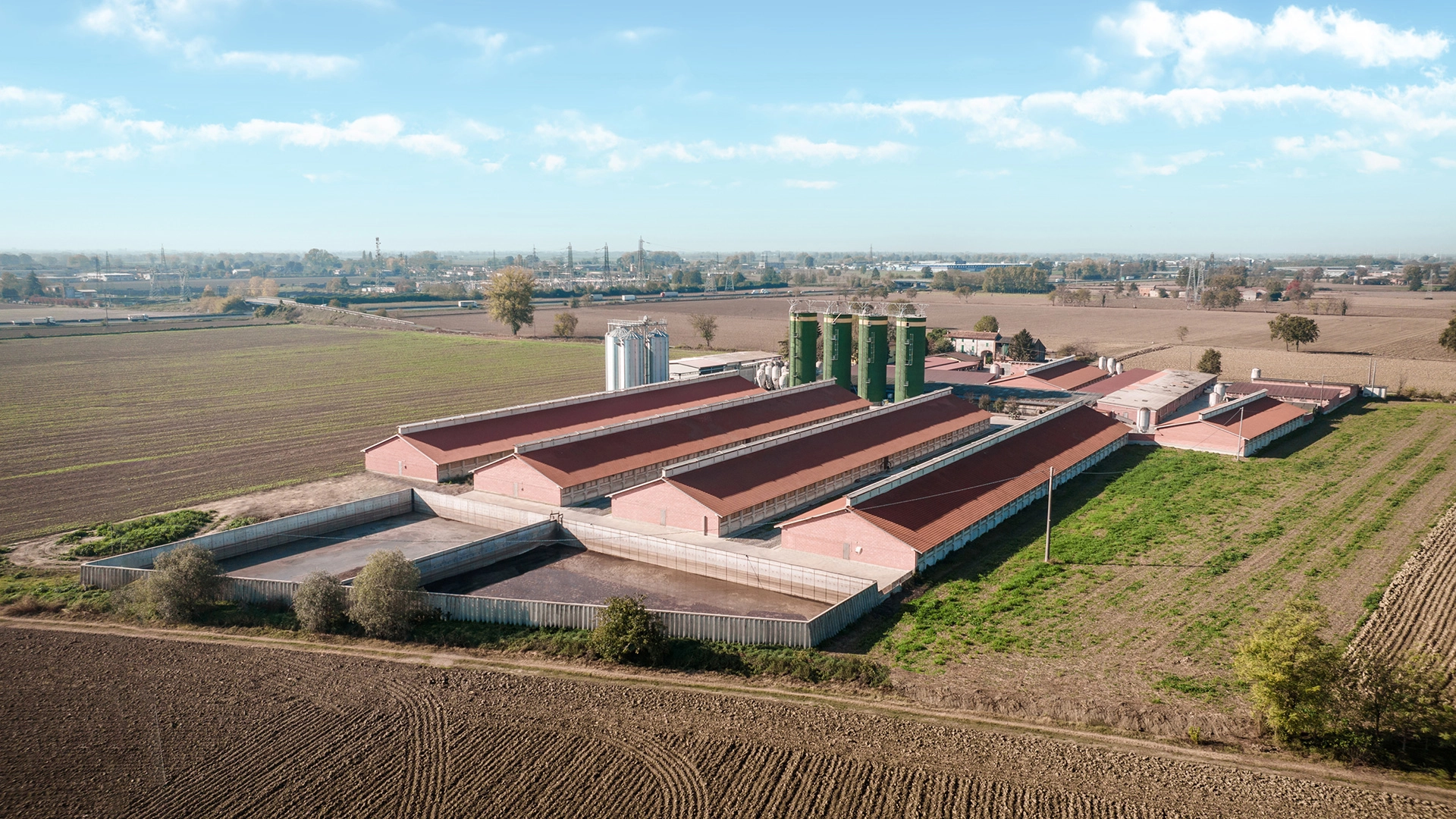Complesso di edifici agricoli con tetti a doppia falda ricoperti da pannelli solari. Le strutture in mattoni rossi sono immerse in un paesaggio rurale, con vasche di raccolta dei liquami.
