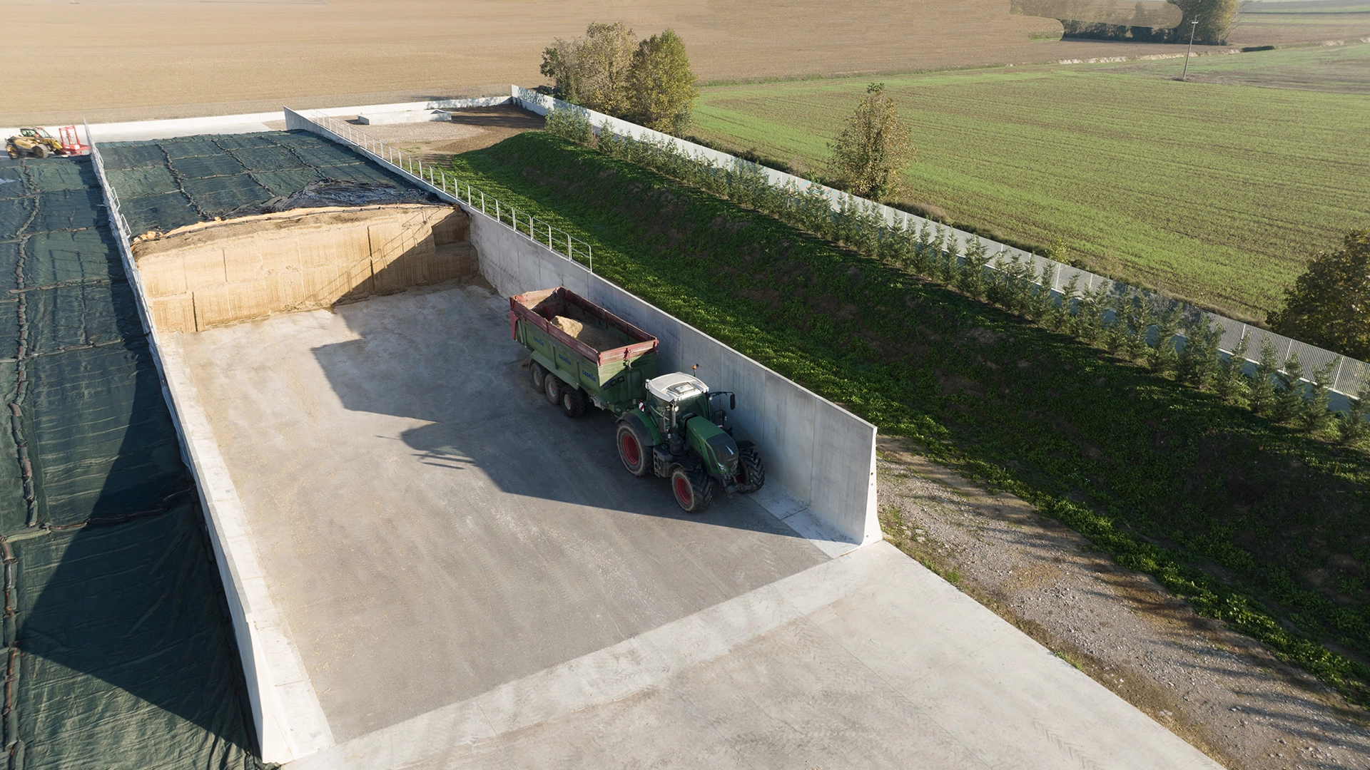 Vista aerea di un'area di stoccaggio agricola con silos in cemento armato prefabbricato. Un trattore con rimorchio è posizionato all'interno di uno dei comparti, parzialmente riempito di foraggio o insilato. A sinistra, un altro comparto è coperto con un telo di protezione.