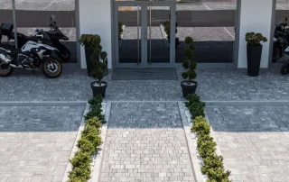 Ingresso con pavimentazione in blocchi grigi, decorato con aiuole di arbusti verdi e ghiaia bianca, affiancato da vasi con piante ornamentali e motociclette parcheggiate vicino alla vetrata d'ingresso.